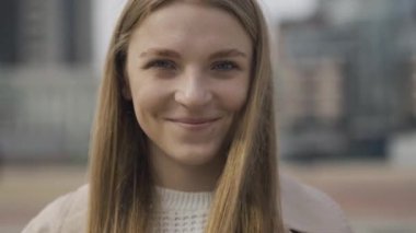 Gri gözlü, kameraya bakıp gülümseyen çekici, beyaz bir kadının yakın çekimi. Bulutlu bir günde şehir meydanında poz veren neşeli, kendine güvenen güzel bir kadının portresi..