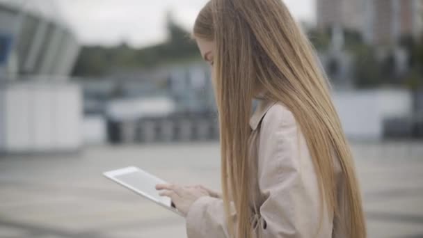 Visão lateral retrato de feliz mulher jovem confiante digitando em tablet, dispositivo de abraço, e sorrindo. Relaxado caucasiano senhora mensagens on-line ou usando mídias sociais ao ar livre em pé na praça urbana da cidade. — Vídeo de Stock