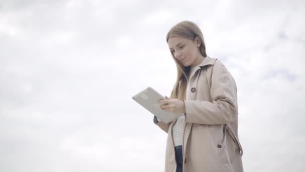Dolní pohled na šťastnou mladou ženu, která používá tablet na pozadí zatažené oblohy, usmívá se a objímá zařízení. Portrét veselé kavkazské studentky nebo podnikatelky zprávy on-line. — Stock video