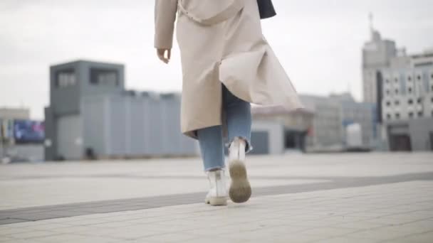 Mulher caucasiana jovem em casaco de primavera ou outono caminhando ao longo da praça da cidade. Visão traseira da senhora confiante em roupas casuais passeando ao ar livre no dia nublado nublado. Conceito de estilo de vida. — Vídeo de Stock