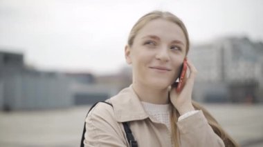 Kamera, akıllı telefondan konuşan beyaz bir kadının güzel yüzünün etrafında dönüyor. Şehir meydanında duran mutlu, rahat, modern bir bayanın yakın plan portresi..