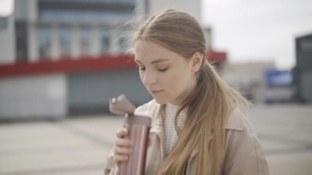 Jovem mulher confiante bebendo café na praça da cidade de garrafa térmica. Retrato de bela senhora caucasiana esperando por alguém ao ar livre no outono nublado ou dia de primavera. Estilo de vida urbano. — Vídeo de Stock