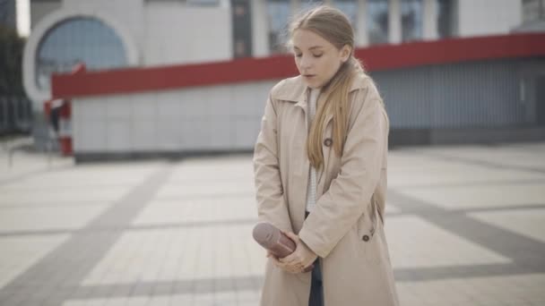 Zamarzająca piękna młoda kobieta stojąca na placu w pochmurny jesienny lub wiosenny dzień i pijąca gorącą kawę lub herbatę z termosu. Portret kobiety rasy kaukaskiej na zewnątrz. — Wideo stockowe