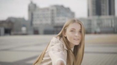 Konseptimi takip et. Genç ve güzel bir kadın kameraya el ele uzanıyor. Şehir meydanında dönüp yürüyoruz. Bulutlu bir günde açık havada eğlenen pozitif gülümseyen beyaz kadın portresi..