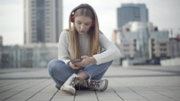 Jeune femme détendue assise sur la place de la ville à écouter de la musique dans les écouteurs et smartphone défilant. Portrait de concentrée belle dame millénaire caucasienne surfant sur les médias sociaux en plein air. — Video