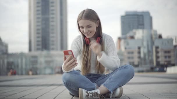 Zrelaksowana kobieta w luźnym stroju siedzi na miejskim placu i macha do smartfona aparatem selfie. Portret beztroskiej białej kobiety korzystającej z czatu wideo na zewnątrz w pochmurny dzień. — Wideo stockowe