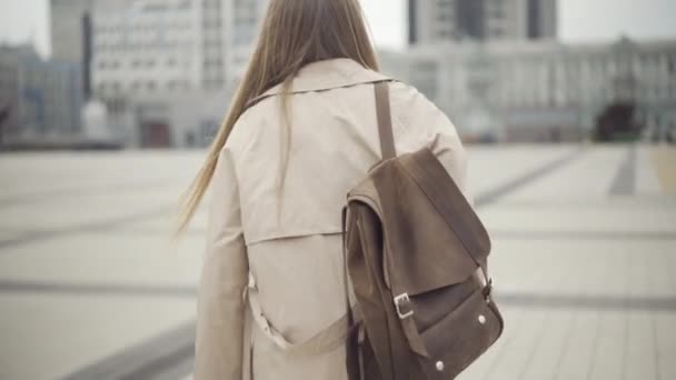 Cámara sigue joven mujer caucásica con mochila caminando por la plaza de la ciudad y mirando a su alrededor. Turista despreocupado o viajero disfrutando de vacaciones en la nublada primavera u otoño ciudad europea. — Vídeo de stock