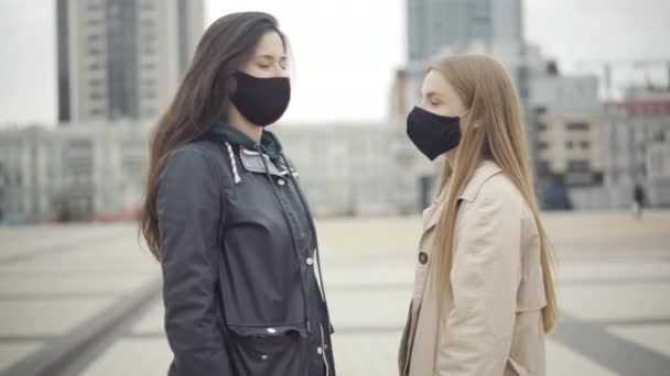 Due giovani donne caucasiche fiduciose in maschere facciali Covid-19 si girano verso la fotocamera e non mostrano alcun gesto con le mani incrociate. Ritratto di amiche in posa all'aperto durante la pandemia di coronavirus. — Video Stock