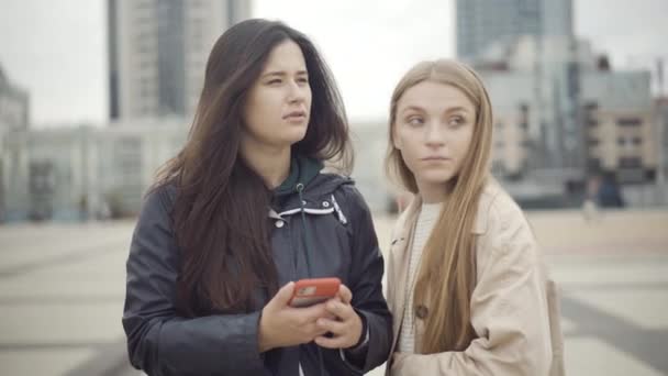 Generade unga turister kontrollera riktning med hjälp av online-karta på smartphone. Porträtt av förlorade vita unga kvinnor som står på torget och pratar. Turism- och GPS-navigeringskoncept. — Stockvideo