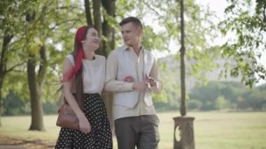 Gülümseyen mutlu sevgili, güneşli parkta yürüyen ve konuşan. Tasasız genç beyaz erkek ve kadının açık havada çıktığı portre. Rahatlamış çift yaz gününün tadını çıkarıyor. Romantizm kavramı.