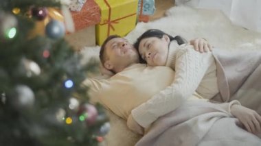 Noel ağacı ve yeni yıl hediyelerinde yumuşak halıda uzanan mutlu, gülen çifte sarılmak. Romantik beyaz erkek ve kadının evde tatilini kutlayan portresi..