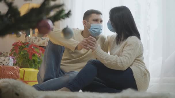 Uvolněný bělošský pár v Covid-19 obličejových maskách sedí doma u novoročního stromku a objímají se. Portrét dospělého muže a ženy oslavující Vánoce na pandemickém uzamčení koronaviru. — Stock video