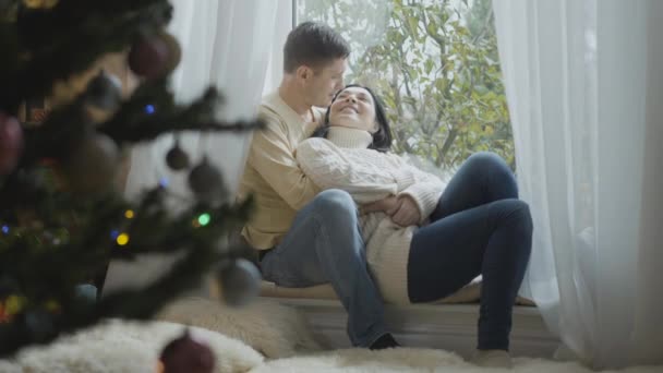 Heureux couple caucasien amoureux assis sur le rebord de la fenêtre le matin de Noël parlant et souriant. Portrait latéral du mari et de la femme positifs profitant des vacances du Nouvel An à la maison. — Video