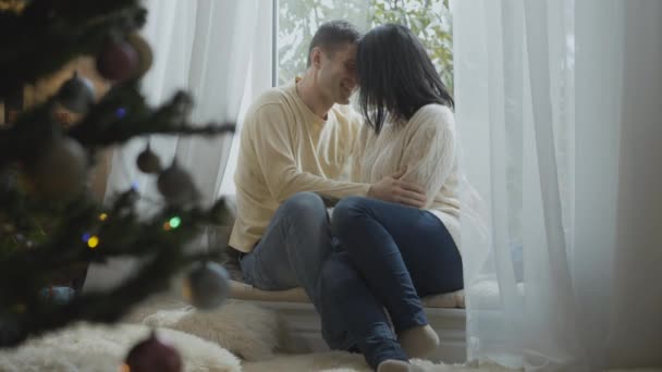 Śmiejąca się kochająca się para siedząca na parapecie twarzą w twarz, rozmawiająca w Nowy Rok. Portret szczęśliwego, zrelaksowanego białego mężczyzny i kobiety korzystających z świąt Bożego Narodzenia w domu. — Wideo stockowe