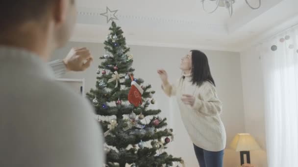 Θετική Καυκάσια γυναίκα στολίζει χριστουγεννιάτικο δέντρο στο σπίτι με έναν θολή άντρα να μιλάει με τη γυναίκα του μπροστά. Πορτρέτο της ευτυχούς κυρίας απολαμβάνοντας διακόσμηση του σπιτιού την παραμονή της Πρωτοχρονιάς. — Αρχείο Βίντεο