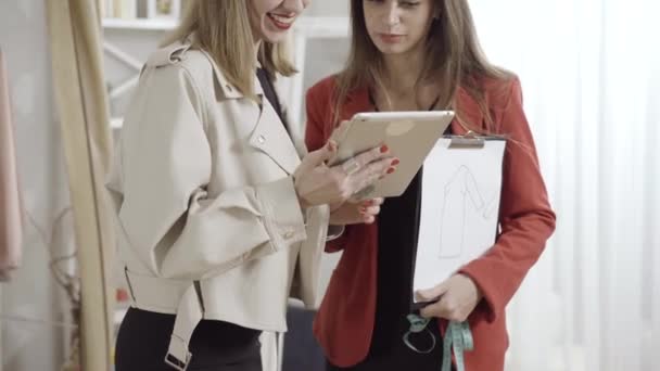 Två oigenkännliga unga leende kvinnor står i ateljé med tablett och klänning design och prata. Positiva kaukasiska designers diskuterar modetrender i workshop. Stil koncept. — Stockvideo