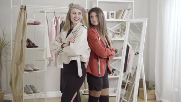 Dos jóvenes mujeres positivas seguras posando espalda con espalda en el taller. Retrato de hermosos sastres o diseñadores caucásicos mirando a la cámara y sonriendo. Concepto de moda y ocupación. — Vídeos de Stock