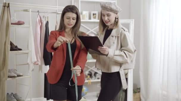 Dos jóvenes diseñadores de moda profesionales que hablan y escriben mediciones en el taller. Retrato de mujeres caucásicas confiadas diseñando un atuendo elegante en interiores. — Vídeos de Stock