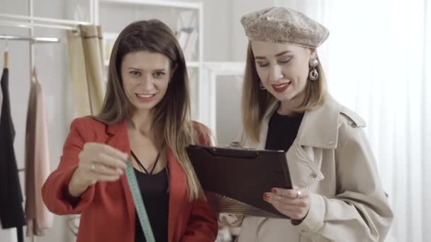 Retrato de un joven diseñador de moda sonriente y sastre usando cinta métrica en el taller. Mujeres caucásicas hermosas confiadas que trabajan en el diseño en interiores hablando. Concepto de moda y belleza. — Vídeos de Stock