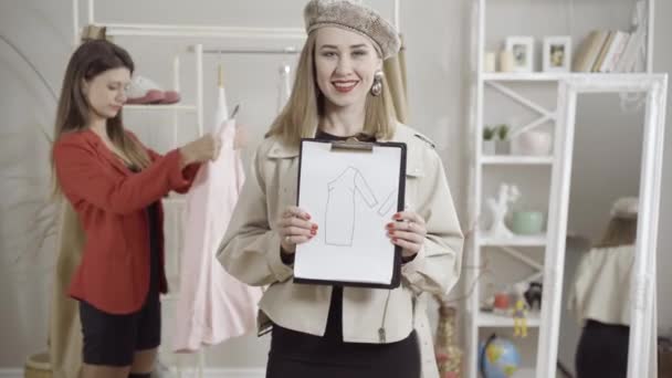 Portrét nádherné mladé ženy pózující s designem šatů v ateliéru jako rozmazaný kolega zkoumající sbírku na pozadí. Pozitivní sebevědomí Kavkazské ženy navrhující módní oblečení. — Stock video