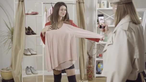 Pozytywna młoda kobieta pozowała ze stylowym różowym pulowerem w atelier jako przyjaciółka robiąca zdjęcia na smartfonie. Szczęśliwy klient kaukaski wybierając koncepcję odzieży wewnątrz. — Wideo stockowe