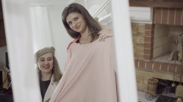 Reflektion i spegeln av nöjd kund undersöker rosa blus i ateljé med positiv designer sömmerska leende i bakgrunden. Unga snygga vita kvinnor väljer plagg inomhus. — Stockvideo