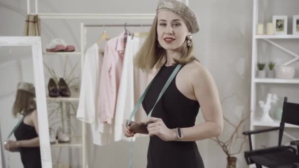 Stylish elegant slim woman in black dress and beret turning to camera and smiling. Portrait of confident positive Caucasian lady posing in atelier with measuring tape. — Stock Video