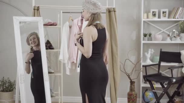 Mujer caucásica delgada feliz admirando la reflexión en el espejo posando en elegante vestido negro y boina. Elegante sastre joven con cinta métrica satisfecho con el nuevo atuendo de moda de pie en el taller. — Vídeos de Stock