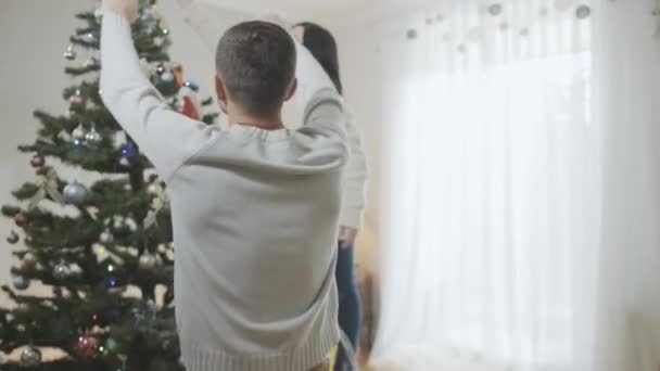 A câmera segue o marido caucasiano ajudando a esposa a decorar a árvore de Natal em casa. Homem caucasiano amoroso que apoia a mulher que decora a árvore de Ano Novo na estação de férias. — Vídeo de Stock