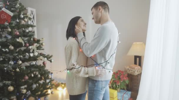 Вид влюбленной пары, стоящей дома у елки лицом к лицу. Счастливый белый мужчина и женщина наслаждаются канун Нового года в помещении. Понятие любви и романтического отдыха. — стоковое видео