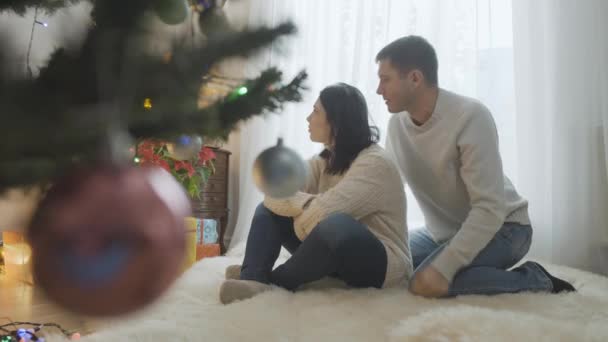 Lächelnder liebevoller Mann im Gespräch mit einer traurigen Frau, die am Weihnachtsbaum sitzt. Aufgebrachte kaukasische Ehefrau stritt an Silvester mit Ehemann. Beziehungsschwierigkeiten in der Ferienzeit. — Stockvideo