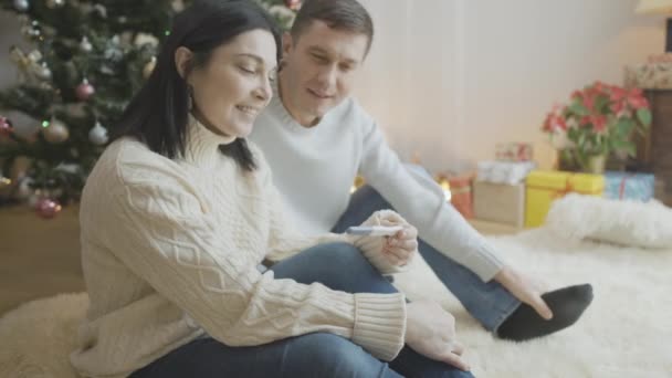 Vista lateral de la mujer embarazada feliz emocionada mirando positivo prueba de embarazo sentado con el hombre sonriente en el árbol de Navidad en casa. Pareja cariñosa caucásica en Año Nuevo en interiores. — Vídeos de Stock