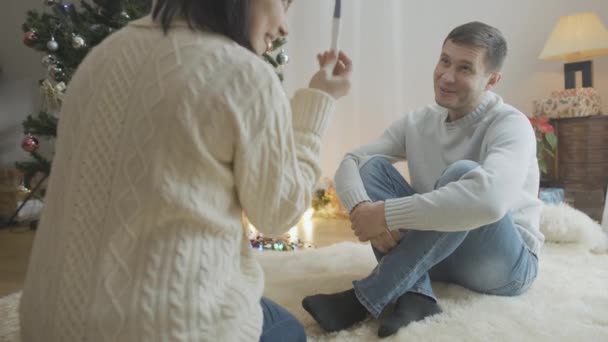 Retrato del hombre caucásico sorprendido sentado en casa en Navidad como mujer que muestra una prueba de embarazo positiva. Marido sorprendido y esposa emocionada celebrando el Año Nuevo juntos en casa. — Vídeos de Stock