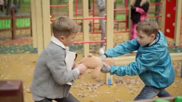 Sonbahar parkında oyun parkında oyuncak için savaşan beyaz çocuklar. İki kardeş oyuncak ayıyı dışarı çekiyor. Çatışma ve çocukluk kavramı. — Stok video
