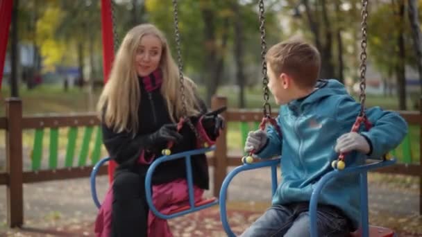 Vreugdevolle ontspannen blanke jongen zittend op schommels praten met moeder en geven high-five. Positieve jongen rust in het weekend met een mooie glimlachende vrouw. Gelukkige familie op speeltuin buiten. — Stockvideo