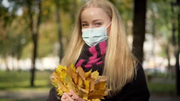 Mooie gelukkige jonge vrouw in Covid-19 gezichtsmasker poseren met gele bladeren boeket in de herfst park. Positieve blanke dame buiten op coronavirus pandemie kijkend naar camera. — Stockvideo