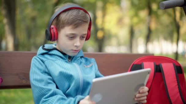 Zaangażowany kaukaski chłopiec w słuchawki gry online na tablecie siedzi w jesiennym parku na świeżym powietrzu. Portret wchłoniętego dziecka cieszącego się wirtualną rzeczywistością w weekendy. Koncepcja uzależnienia od gier wideo. — Wideo stockowe