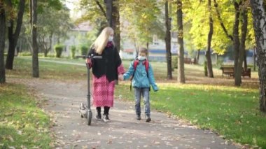 Geniş açıda, Coronavirus maskeli Kafkas anne ve oğlu elektrikli scooterla sonbahar parkında yürüyorlar ve konuşuyorlar. Genç bir kadın ve yakışıklı bir çocuğun portresi. Salgın bir şekilde dışarıda geziniyor..