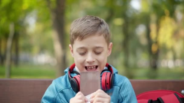 Çikolata yiyen ve lezzetli bir tatlı çiğneyen mutlu, rahat bir çocuğun yakın plan portresi. Neşeli şirin beyaz çocuk dışarıda lezzetli yemekler yiyor sonbahar parkında bankta oturuyor.. — Stok video