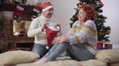 Neşeli Kafkasyalı adam gülümseyen neşeli kadının kafasına Noel şapkası takıyor. Olumlu bir çift yeni yıl arifesinde evde eğleniyor. Noel kutlaması ve neşe.