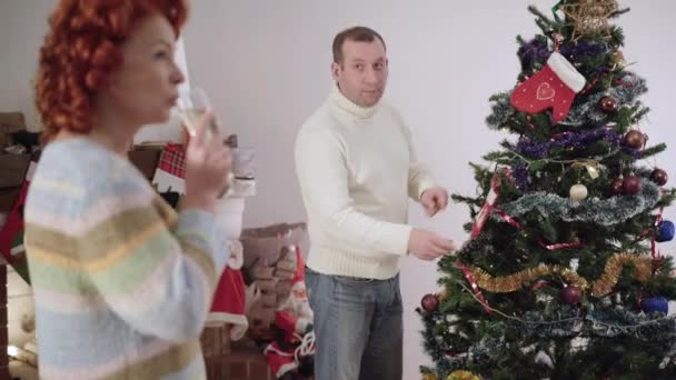 一个紧张的白人男人装饰圣诞树的画像，就像一个模糊的女人在前面喝香槟而忽略了丈夫。在室内准备庆祝新年的一对心烦意乱的成年夫妇. — 图库视频影像
