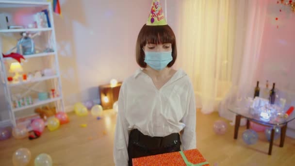 Vidám fiatal kaukázusi nő Covid-19-es maszkban és parti sapkában ajándékdobozokat kap otthon születésnapjára. Boldog izgatott gyönyörű hölgy ünnepli nyaralás koronavírus világjárvány lezárása. — Stock videók