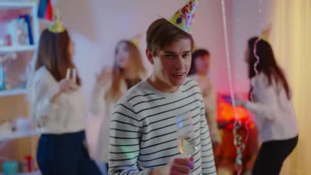 Portrait d'un bel homme millénariste souriant cliquetant un verre à vin avec appareil photo, buvant de l'alcool et dansant avec des amis flous s'amusant en arrière-plan. positif caucasien gars célébrer anniversaire. — Video