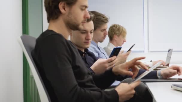 Vista lateral de quatro jovens caucasianos concentrados sentados no cargo usando gadgets e pensamento. Colegas planejando inicialização na sala de conferências dentro de casa. Conceito de negócio e trabalho em equipe. — Vídeo de Stock