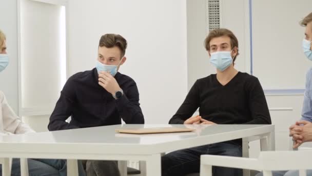 Des hommes blancs concentrés du millénaire portant des masques Covid assis à la table de conférence font du remue-méninges avec leurs collègues. Des jeunes entrepreneurs ciblés envisageant de démarrer une entreprise contre la pandémie de coronavirus. — Video