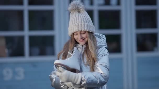 Joyeux mignon fille caucasienne tenant patins à glace regardant la caméra et souriant. Portrait d'un joli enfant joyeux posant à l'extérieur sur une patinoire. Concept sport et enfance. — Video