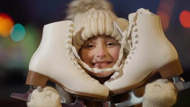 Opgewonden gelukkig blank meisje met nieuwe schaatsen die glimlachend naar de camera kijken. Close-up headshot van vrolijk kind ontvangen geschenk voor Nieuwjaar met kerstverlichting op de achtergrond in de nacht stad. — Stockvideo