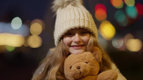 Gros plan portrait d'une jolie fille heureuse posant avec un ours en peluche à l'extérieur la nuit de Noël. Charmante enfant caucasien souriant tenant jouet et regardant la caméra avec des lumières clignotant à l'arrière-plan. — Video
