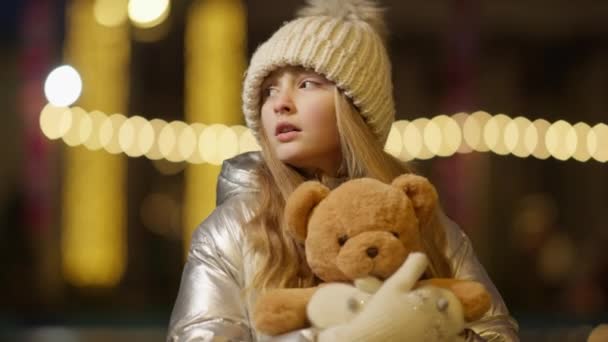 Portret smutnej dziewczyny zagubionej w miejskim mieście w świąteczną noc. Stresowany biały dzieciak przytulający pluszowego misia rozglądającego się ze złotymi girlandami w tle. — Wideo stockowe