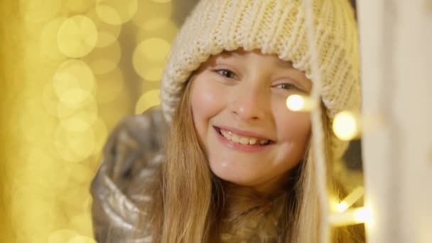 Cara de niña feliz sonriente de pie al aire libre en luces doradas de Navidad y mirando a la cámara. Retrato en la cabeza del encantador niño caucásico alegre posando en Año Nuevo por la noche. — Vídeos de Stock